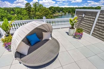 A white wicker chair with a blue pillow is on a balcony with a view of a lake and trees.